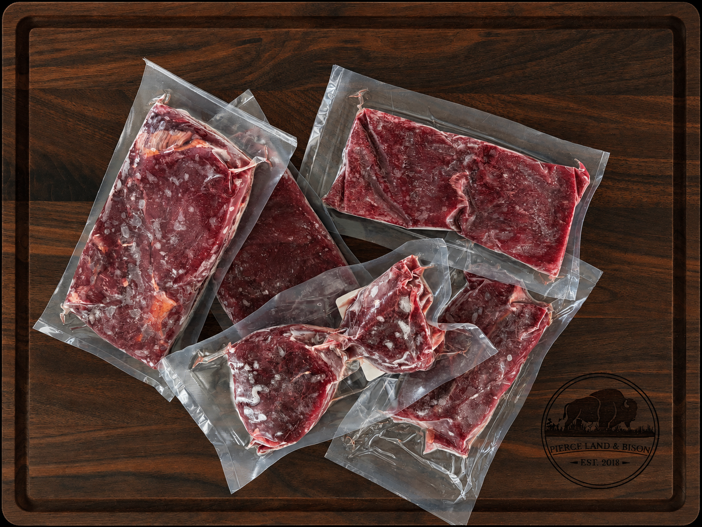 The Steak Sampler from Pierce Land & Bison showing five vacuum-sealed packages of grass-fed bison steaks including flat iron, top sirloin, filet mignon, New York strip, and ribeye on a dark walnut cutting board